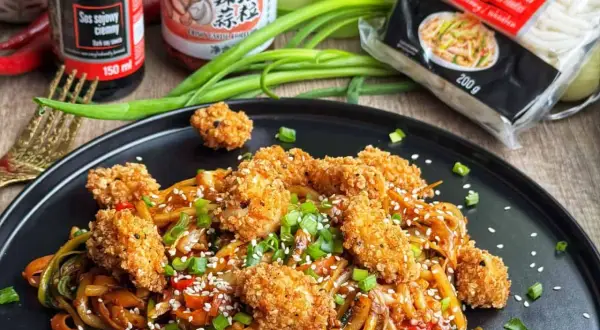 Pyszne udon z kurczakiem w panierce, posypane sezamem i szczypiorkiem. Obok sos sojowy i opakowanie makaronu udon.