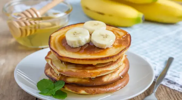 En stapel gyllene bananpannkakor toppas med bananskivor och ringlad honung. Ett enkelt bananpannkaka recept för en härlig frukost.