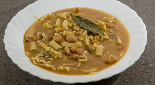 Un piatto fumante di pasta e fagioli, con pasta corta, fagioli borlotti, carote e una foglia di alloro.