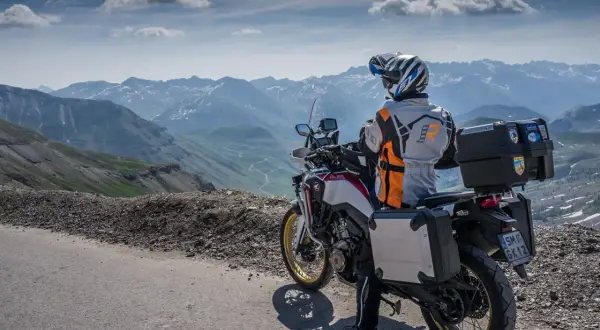 Motocyklista w kasku i kurtce podziwia górską panoramę Alp z krętą drogą w dole.