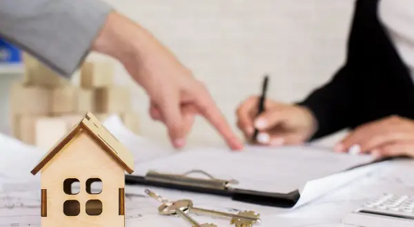 Un perito per valutazione casa indica un documento mentre un'altra persona firma. Chiavi e modellino di casa sul tavolo.