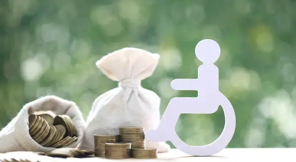 Symbol osoby niepełnosprawnej obok worków z pieniędzmi i monet. Finansowe wsparcie dla osób z niepełnosprawnością.