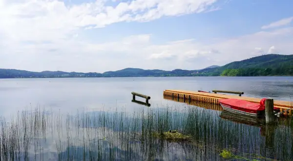 Ein Steg mit einem roten Boot am Laacher See. Schilf ragt aus dem ruhigen Wasser, das die bewaldeten Hügel am Horizont spiegelt.