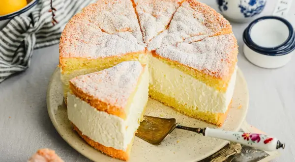 Saftige Quark Sahne Torte, mit Puderzucker bestäubt, auf einem Teller mit Tortenheber. Daneben Zitronen und eine Kanne.