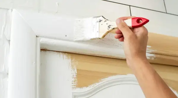 Mano che dipinge con un pennello bianco una porta interna in legno, rinnovando l'aspetto.