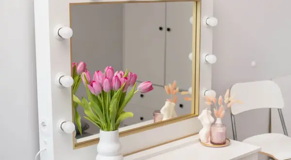 Stylowe lustro z lampkami, idealne do makijażu. Obok bukiet różowych tulipanów w wazonie.