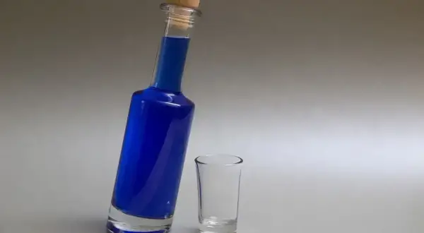 Niebieska wódka w butelce z korkiem i pusty kieliszek obok.