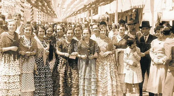 Grupo de mujeres y niñas con trajes de volantes y lunares, evocando el origen del traje de flamenca en una feria antigua.