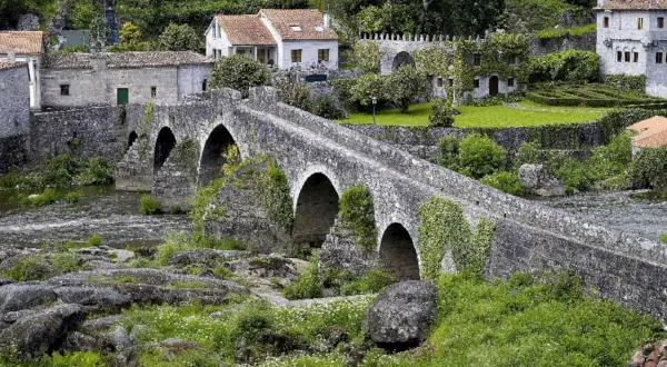 Un antiguo ponte carreira de piedra cruza un río, rodeado de casas rústicas y vegetación exuberante.