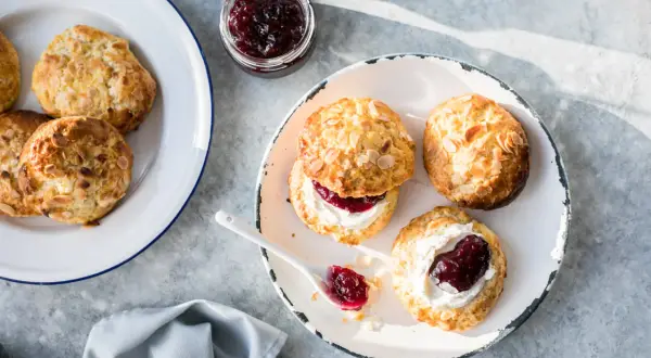 Nybakade scones med mandelflagor, grädde och sylt på en tallrik. En härlig fika!