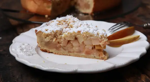 Ein Stück apfelkuchen sehr fein, mit Streuseln und Puderzucker bestäubt, auf einem weißen Teller mit Gabel und Apfelscheiben.