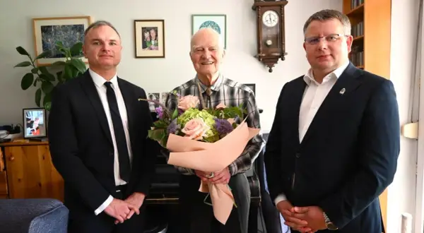 Trzech mężczyzn, jeden z bukietem kwiatów, w eleganckich strojach.