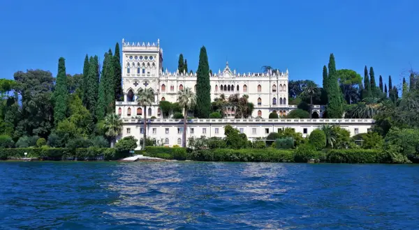 Villa neogotica su un'isola del Lago di Garda, circondata da cipressi e vegetazione lussureggiante, con il blu intenso dell'acqua in primo piano.