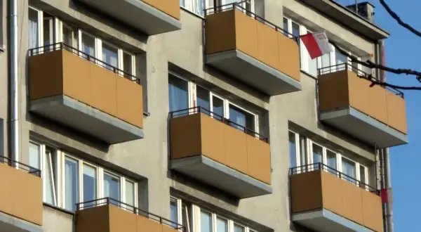 Szare bloki z brązowymi balkonami, typowe dla mieszkania komunalnego. Widać polską flagę.