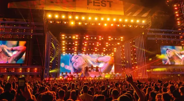 Tłum na festiwalu Fest, rozświetlony sceną i ekranami z artystami. Widać logo "FEST festival" i tłum ludzi z telefonami.