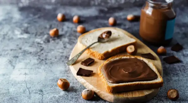 Tranche de pain généreusement tartinée de pâte à tartiner maison 3 ingrédients. Des noisettes et des carrés de chocolat parsèment la table.