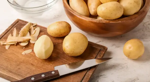 Vorbereitung für Kartoffelgerichte: Ein Messer liegt auf einem Holzbrett neben geschälten und ungeschälten Kartoffeln. Eine Schüssel mit Kartoffeln steht daneben. Wie viel Kartoffeln pro Person?