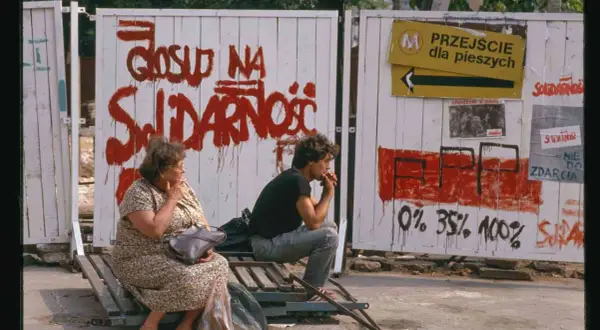 Dwie osoby siedzą na ławce przed płotem z napisem "Głosuj na Solidarność". To symboliczny obraz upadku komunizmu.