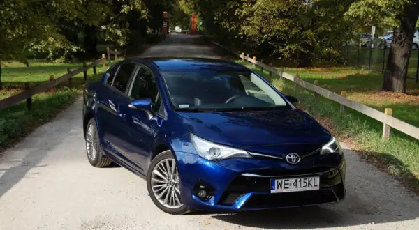 Błękitna Toyota Avensis T27 na żwirowej drodze, otoczona zielenią i drzewami.