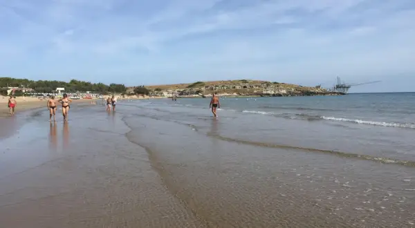 Persone passeggiano sulla battigia di Cala Molinella, con un trabucco sullo sfondo.