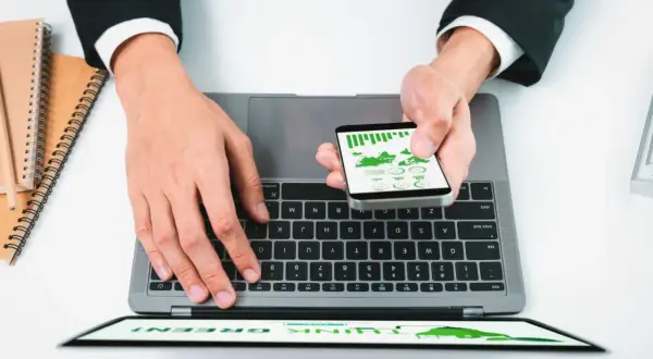 Praca przez internet: dłonie na klawiaturze laptopa, smartfon z wykresami w ręce, notatniki obok.