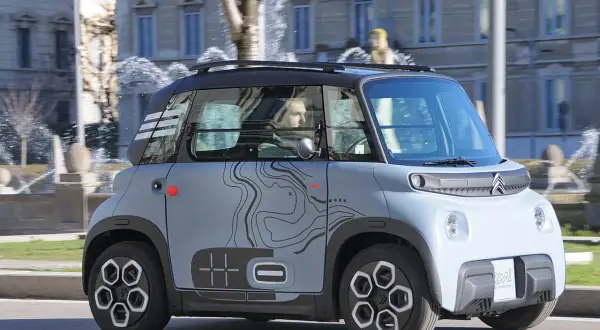 Citroën Ami, un veicolo elettrico compatto, sfreccia con la sua velocità massima in città, con uno sfondo di fontane e palazzi.