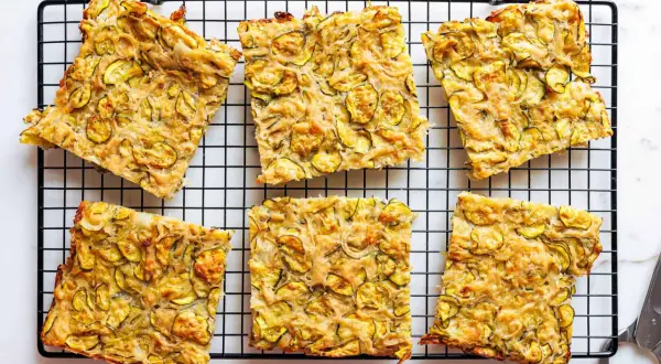 Sechs rechteckige Stücke einer gebackenen Zucchinipfanne mit gelben Zucchinischeiben auf einem schwarzen Gitter.