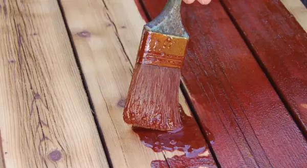 Application d'une teinture rouge sur des planches de bois. Un pinceau chargé de produit montre comment peindre du bois pour le protéger.