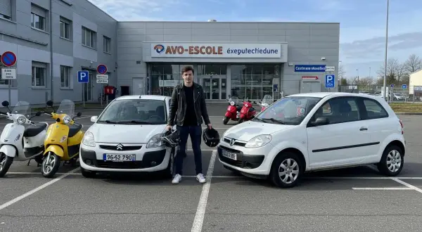 Un jeune homme pose devant une auto-école avec deux voitures sans permis et des scooters. Il tient deux casques, prêt pour son apprentissage.