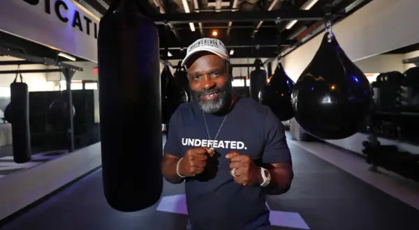 A man in a "UNDEFATED" shirt and Mayweather Boxing + Fitness hat poses with fists up in a gym, ready for Floyd Mayweather training.