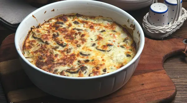 Herzhafter Kohlrabi-Auflauf mit Pilzen und Käse überbacken, serviert in einer weißen Auflaufform auf einem Holzbrett.