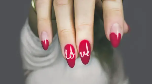 Czerwone paznokcie z białym napisem "love" i zdobieniem w kształcie serduszka.
