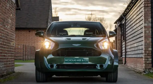 Aston Martin Cygnet w ciemnozielonym kolorze, z reflektorami świecącymi w stronę widza, zaparkowany na podjeździe między ceglanymi budynkami.
