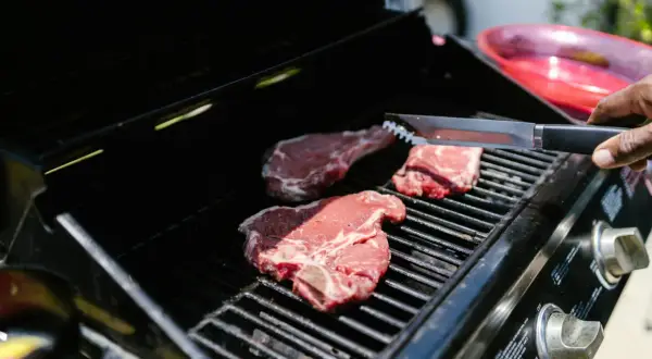 Trzy soczyste steki na ruszcie grilla, gotowe do grillowania. Dłoń z szczypcami zbliża się do mięsa.