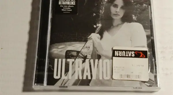 Okładka płyty Lana Del Rey Ultraviolence. Na czarno-białym zdjęciu artystka w białej koszulce, obok naklejka Saturn z ceną.