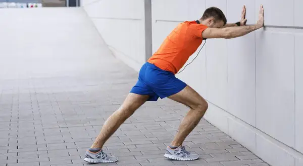 Un homme en tenue de sport fait un étirement mollet contre un mur blanc.