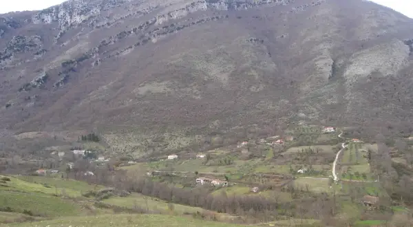 Un paesaggio collinare dove il Cilento mostra case sparse tra alberi spogli e prati verdi, sotto un imponente monte roccioso.