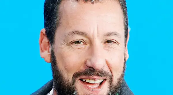 Uśmiechnięty Adam Sandler, gwiazda wielu komediowych filmów, na tle niebieskiego tła.