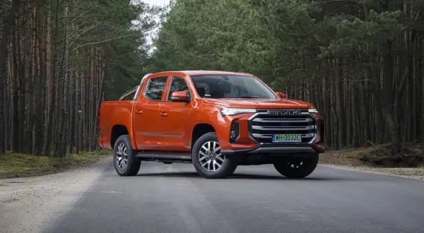 Pomarańczowy Maxus T90 na leśnej drodze. Ten potężny pickup z charakterystycznym grillem i nowoczesnymi światłami świetnie prezentuje się w otoczeniu drzew.