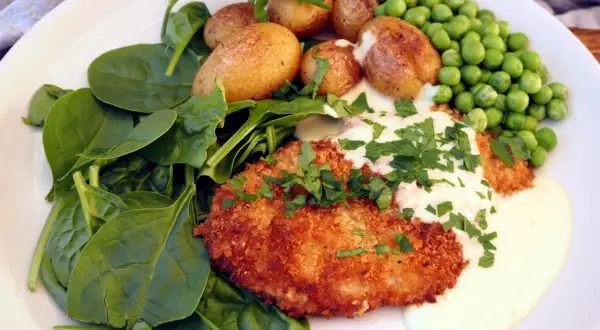 En panerad schnitzel med krämig sås, serverad med färsk spenat, små potatisar och gröna ärtor. En klassisk rätt där såsen till schnitzel lyfter hela smakupplevelsen.