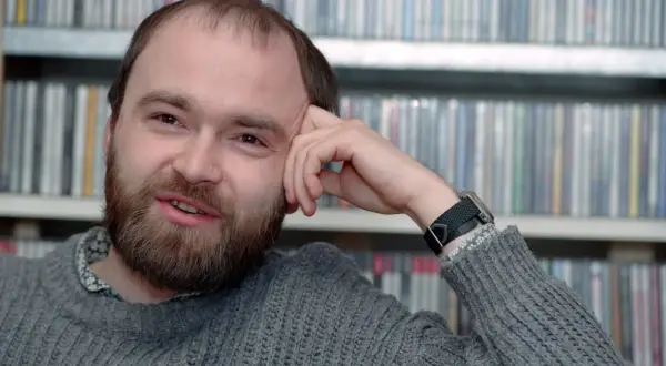 Tomasz Beksiński, zamyślony, z ręką opartą o czoło, na tle regałów z płytami. Jego spojrzenie zdaje się wybiegać poza kadr, jakby rozmyślał o przemijaniu i śmierci.