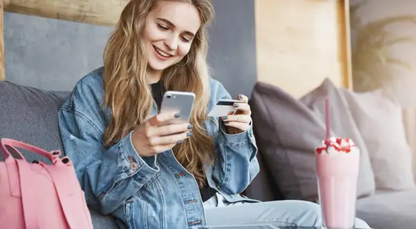 Uśmiechnięta kobieta w dżinsowej kurtce trzyma telefon i kartę płatniczą.