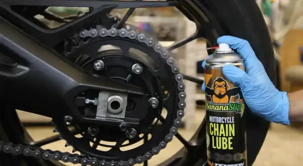 Ręka w niebieskiej rękawiczce aplikuje smar do łańcucha motocyklowego z puszki Banana Slip.
