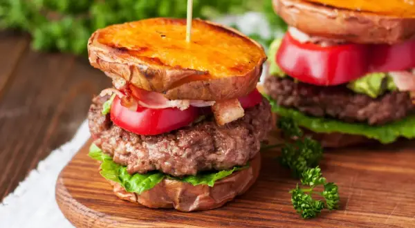 Pyszny burger na talerzu z soczystą wołowiną, chrupiącym bekonem, pomidorem i awokado, podany na słodkich ziemniakach.