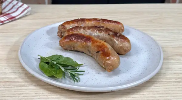 Tre salsicce dorate e succulente, appena cotte in padella, riposano su un piatto con rosmarino e alloro.