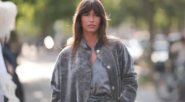 Frau mit curtain bangs und natürlichen Locken trägt einen grauen Kunstpelzmantel und grüne Hose.