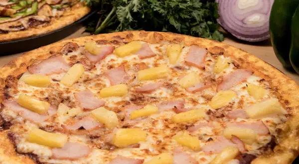 Pizza hawajska składniki: kawałki ananasa i szynki na serze. W tle inna pizza i warzywa.