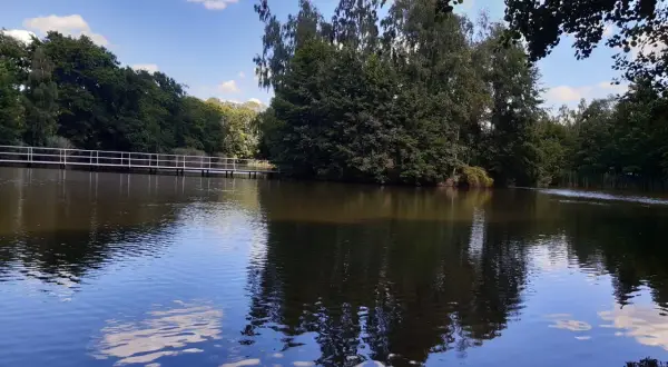 Spokojne jezioro z odbijającymi się chmurami i drzewami. W oddali biały mostek, idealne miejsce na wypoczynek w Golina ośrodek wypoczynkowy.