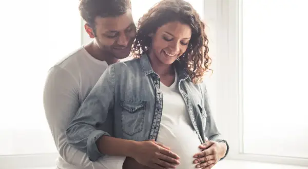 Un couple heureux, le futur papa enlace tendrement le ventre arrondi de sa compagne. Ils pensent déjà au prénom en N pour leur bébé.