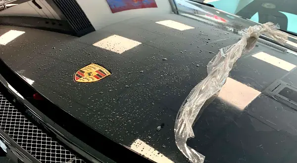 Czarna maska Porsche z logo, na której widać krople wody i fragment folii. Trwa klejenie folii na sucho.